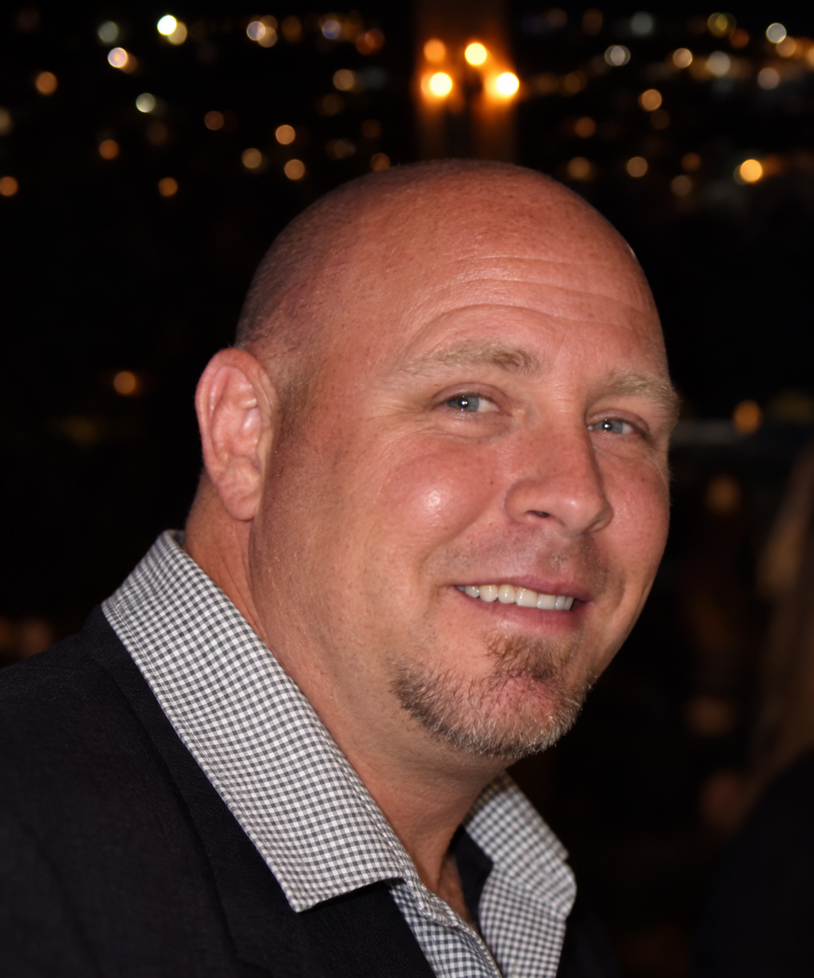 Ron Devine smiling in a checked shirt and dark jacket, with warm lights behind him